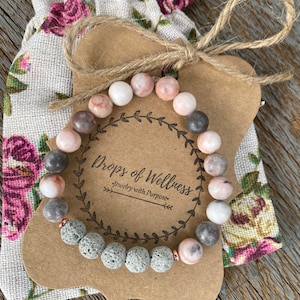May include: A gray and pink beaded bracelet with lava rock beads and gold accents. The bracelet is displayed on a brown card with the text "Drops of Wellness Jewelry with Purpose".