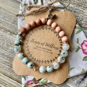 以下が含まれることがあります： 緑と白の斑点のある石のブレスレットに、ローズゴールドのハートチャームと木製のビーズが付いています。ブレスレットは、小さな花の布製の袋に詰められており、茶色の紙のタグには「Drops of Wellness Jewelry with Purpose」と書いてあります。