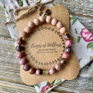 May include: A pink and brown gemstone bracelet with a gold heart charm. The bracelet is on a brown card with the text "Drops of Wellness Jewelry with Purpose".