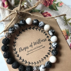 May include: A black lava rock and white and black marbled stone bracelet with silver beads. The bracelet is on a brown card with the text "Drops of Wellness - Jewelry with Purpose".