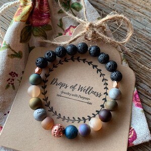 May include: A beaded bracelet with black lava rock beads and various colorful stone beads. The bracelet is displayed on a brown tag with the text "Drops of Wellness - jewelry with Purpose". The bracelet is presented with a floral drawstring bag.
