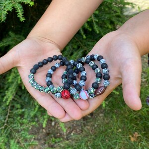 May include: Four black lava stone bracelets with different sports-themed charms. The charms include a football, baseball, and basketball.