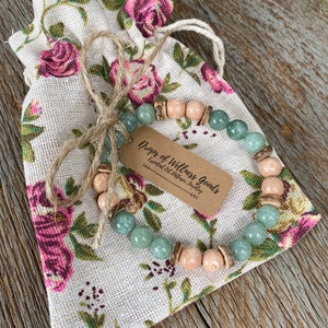 Può includere: Un braccialetto con perline verdi e marroni con un piccolo cartellino marrone che recita "Drops of Wellness Goods - Essential Oil Diffuser Jewelry". Il braccialetto è in una piccola borsa a cordoncino floreale.