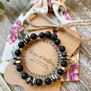 May include: Black and white beaded bracelet with gold accents and lava rock beads. The bracelet is displayed on a brown card with the text "Drops of Wellness Jewelry with Purpose".