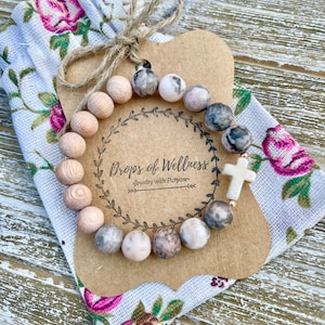 May include: A beaded bracelet with a cross charm rests on a floral fabric pouch and a brown paper tag. The bracelet features wooden beads and patterned stone beads. The tag reads "Drops of Wellness - Jewelry with Purpose."