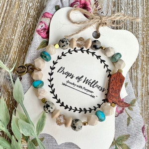 May include: A brown and white speckled stone bracelet with turquoise and gold accents. The bracelet is on a white card with the text "Drops of Wellness Jewelry with Purpose".