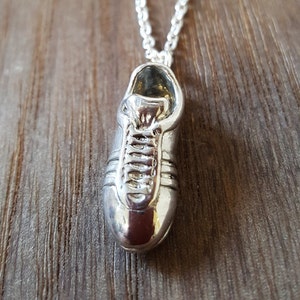 May include: A silver pendant necklace featuring a detailed, three-dimensional shoe charm. The shoe is silver and has a lace-up design.