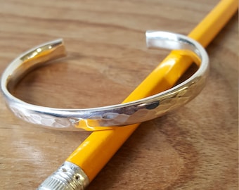 May include: A silver hammered bangle bracelet. The bracelet is open and is resting on a yellow pencil.