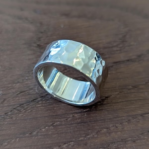 May include: A wide, silver ring with a hammered texture. The ring has a flat band and a slightly curved surface, reflecting light. The ring is displayed on a dark brown wooden surface.