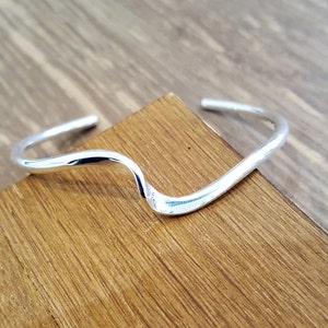 May include: A silver-colored cuff bracelet with a wave design. The bracelet is open at the ends and sits on a wooden surface. The bracelet's smooth, curved lines create a minimalist and modern aesthetic.