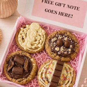 May include: A white gift box containing four large cookies topped with various chocolates and sweets, nestled on pink shredded paper. The inside lid features a pink rectangular label with the black text: FREE GIFT NOTE GOES HERE!