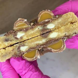 May include: A split cookie featuring a golden-brown dough base embedded with white chocolate chips and pieces of milk chocolate candy filled with a smooth, amber-colored caramel center, revealing a soft, textured interior and a rich, layered appearance.