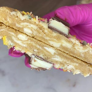 May include: A split cookie featuring a golden-brown dough base embedded with white chocolate chunks and pieces of milk chocolate candy. The surface is decorated with colorful sprinkles, and the interior reveals a soft, dense texture throughout.