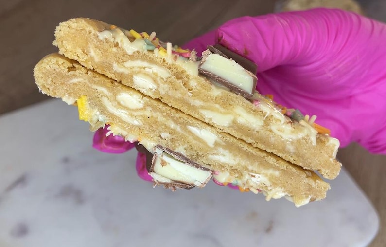 May include: A split cookie featuring a golden-brown dough base embedded with white chocolate chunks and pieces of milk chocolate candy. The surface is decorated with colorful sprinkles, and the interior reveals a soft, dense texture throughout.