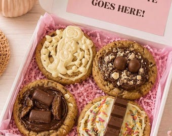 May include: A white gift box containing four large round cookies topped with various chocolate candies, including white chocolate drizzle, chocolate spheres, Cadbury chocolate pieces, and a chocolate bar with colorful sprinkles on pink paper.