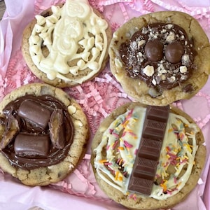 May include: Four round cookies with various toppings. One has white chocolate drizzle and a figure, one has milk chocolate spread with round sweets, one has chocolate spread with caramel candy pieces, and one has white drizzle, sprinkles, and a chocolate bar.