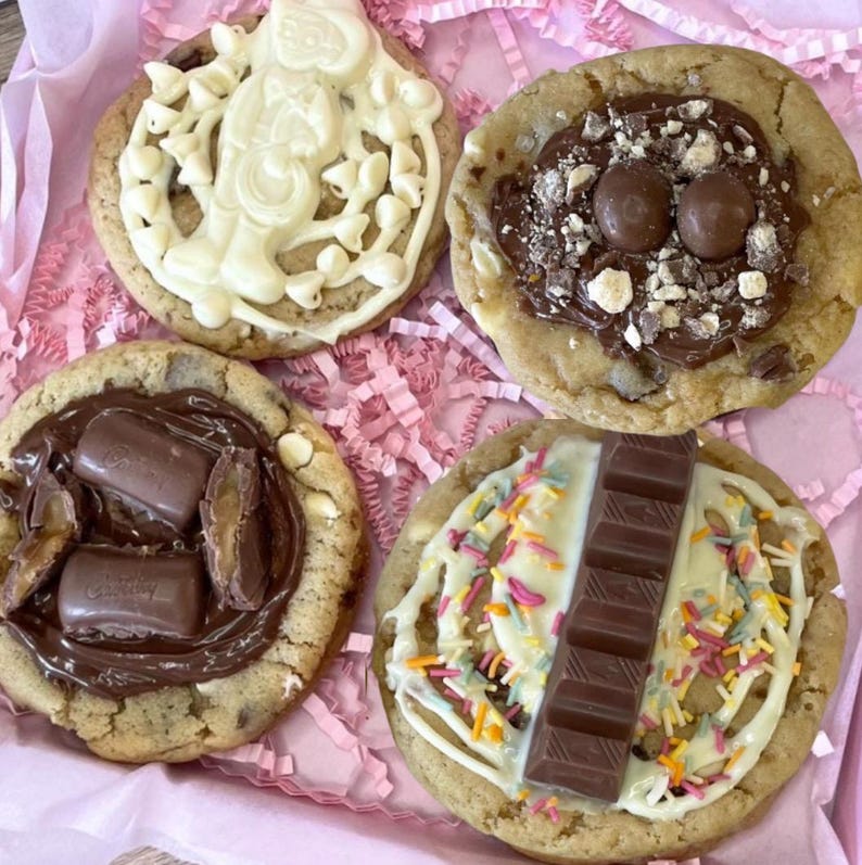 May include: Four round cookies with various toppings. One has white chocolate drizzle and a figure, one has milk chocolate spread with round sweets, one has chocolate spread with caramel candy pieces, and one has white drizzle, sprinkles, and a chocolate bar.