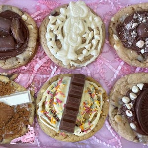 May include: A collection of six round cookies topped with various chocolate confections, including chocolate bars, white chocolate drizzle, sprinkles, and crushed biscuits, arranged in a grid on a light pink shredded paper background.