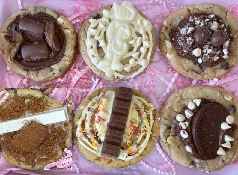 May include: A collection of six round cookies topped with various chocolate confections, including chocolate bars, white chocolate drizzle, sprinkles, and crushed biscuits, arranged in a grid on a light pink shredded paper background.