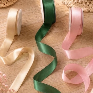 May include: Three spools of satin ribbon in cream, dark green, and light pink. Each spool features a smooth, shiny finish and is partially unrolled, with the ribbons extending across the surface in gentle, flowing curves and soft, elegant folds.
