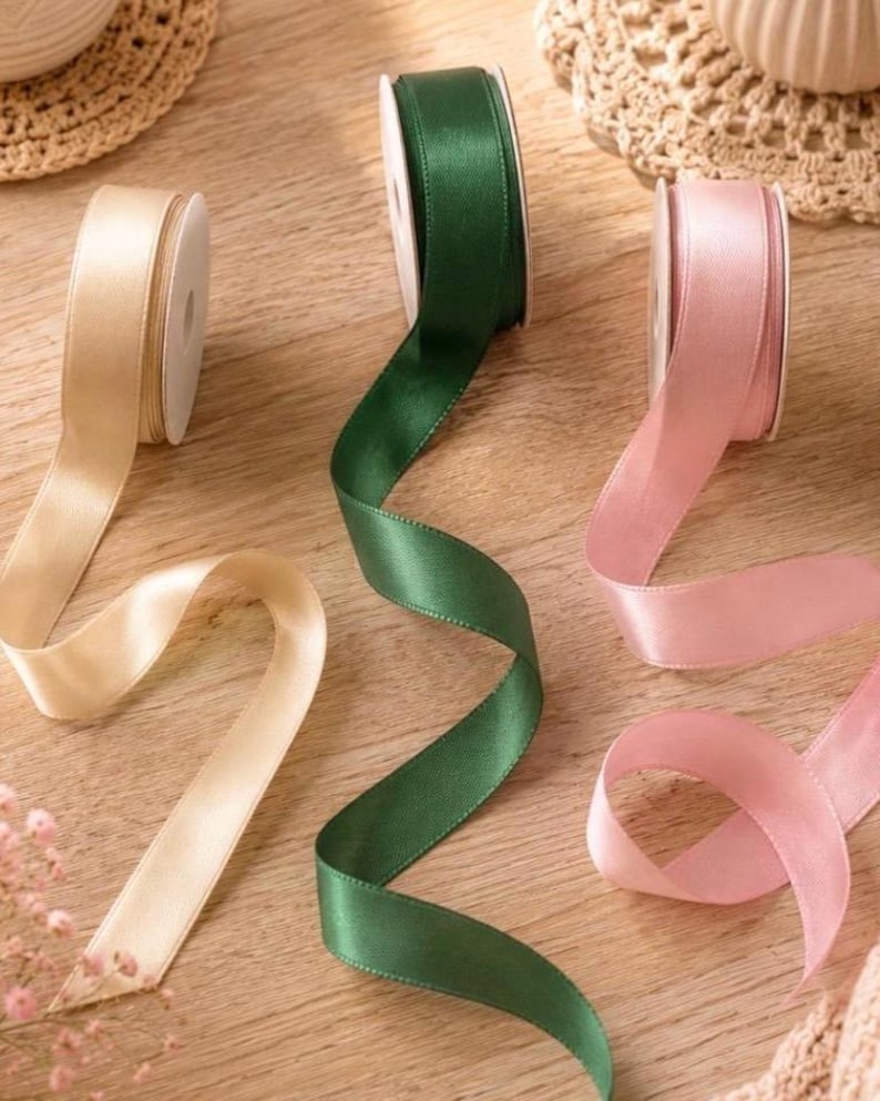 May include: Three spools of satin ribbon in cream, dark green, and light pink. Each spool features a smooth, shiny finish and is partially unrolled, with the ribbons extending across the surface in gentle, flowing curves and soft, elegant folds.