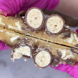 May include: A golden brown cookie broken in half, revealing a soft interior with white chocolate chips and embedded pieces of chocolate-covered malted milk balls. The cookie dough has a textured surface with visible chocolate and malted candy pieces.