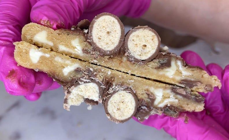 May include: A golden brown cookie broken in half, revealing a soft interior with white chocolate chips and embedded pieces of chocolate-covered malted milk balls. The cookie dough has a textured surface with visible chocolate and malted candy pieces.