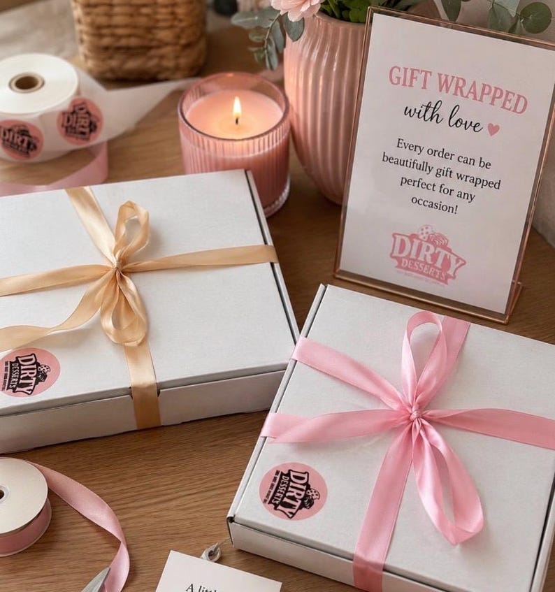 May include: Two square white cardboard boxes tied with satin ribbons, one in champagne and one in light pink. Each box features a round pink sticker with the text Dirty Desserts. A sign reads GIFT WRAPPED with love and Dirty Desserts.