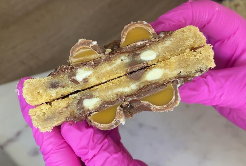May include: A split cookie featuring a golden-brown dough base embedded with white chocolate chips and pieces of milk chocolate candy filled with a smooth, amber-coloured caramel centre, revealing a soft, textured interior and a rich, layered appearance.