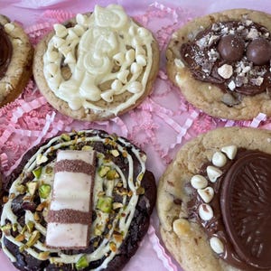 May include: Five round cookies with various toppings. One has chocolate bars, one has white chocolate drizzle and chips, one has chocolate spheres and crumbles, one has shredded topping, and one has a chocolate wafer, pistachios, and white chocolate chips.