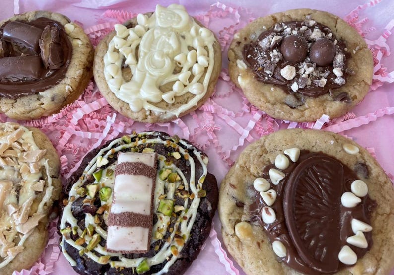 May include: Five round cookies with various toppings. One has chocolate bars, one has white chocolate drizzle and chips, one has chocolate spheres and crumbles, one has shredded topping, and one has a chocolate wafer, pistachios, and white chocolate chips.
