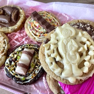 May include: A collection of six round cookies with various toppings including chocolate bars, white chocolate chips, sprinkles, and chopped nuts. One cookie features a white chocolate figure, while another has a small chocolate bar with visible text.