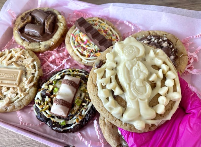 May include: A collection of six round cookies with various toppings including chocolate bars, white chocolate chips, sprinkles, and chopped nuts. One cookie features a white chocolate figure, while another has a small chocolate bar with visible text.