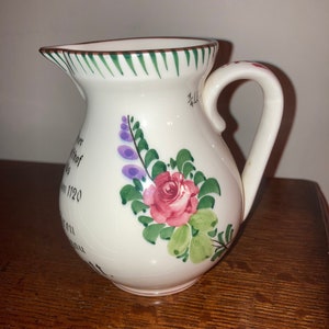 May include: White ceramic pitcher with a green and brown striped rim. The pitcher has a floral design with a pink rose, green leaves, and purple flowers. The pitcher has a handwritten inscription on the side.