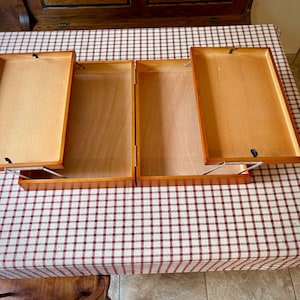 May include: A wooden storage box with two hinged, lift-up lids that open outwards. The box features a light brown wood grain finish, rectangular compartments, and silver-coloured metal support arms attached to the underside of each lid.