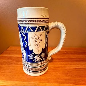Peut inclure: Une chope à bière en céramique avec un corps bleu profond et des détails en relief blancs, incluant une bannière centrale, des motifs floraux et une anse latérale. Le bord porte l'inscription 0.5L, indiquant sa capacité, et la base est ornée.