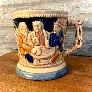 May include: A ceramic mug featuring a raised, three-dimensional relief scene of three figures seated at a table. The mug has a cream-colored base, a blue bottom rim, and a handle textured to resemble a tree branch with bark-like details.