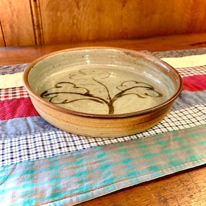 May include: A shallow, round ceramic bowl with a speckled light gray interior and a dark brown rim. The center of the bowl features a hand-painted, dark brown line drawing of a stylized plant with three distinct, rounded leaf clusters.