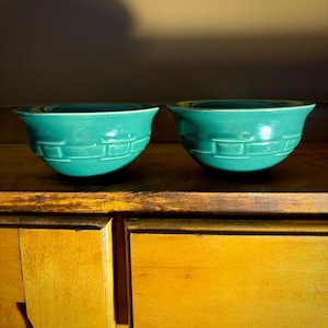 May include: Two turquoise ceramic bowls with a geometric pattern. The bowls sit on a wooden surface. Each bowl has a flared rim. The bowls are a matching set and are likely vintage. The bowls are a teal colour.