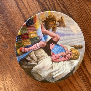 May include: A ceramic plate with a colorful patchwork quilt design. A young child is lying on a bed, reading a book titled "Dream Big" with a teddy bear beside them.