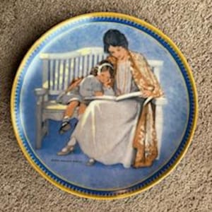 Puede incluir: Un plato de porcelana azul y blanco con una pintura de una madre y un niño leyendo un libro en un banco. La madre lleva una chal marrón y el niño lleva un vestido blanco.