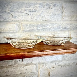 Puede incluir: Dos fuentes de vidrio transparente con un diseño geométrico texturizado. Las fuentes de forma ovalada tienen un borde ligeramente ensanchado. Se exhiben en un estante de madera contra una pared de ladrillos blancos. Las fuentes son probablemente para servir aperitivos o porciones pequeñas.