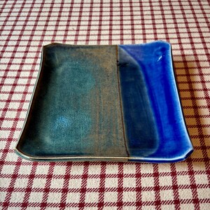 May include: A square ceramic plate featuring a two-tone glaze design. The left side displays a muted teal and brown mottled finish, while the right side is finished in a vibrant, glossy cobalt blue. The plate has slightly raised, curved edges.