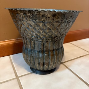 May include: A decorative metal vase with a distressed, aged appearance. The vase has a wide, fluted top with a floral design, a diamond pattern on the body, and a rounded base. The vase is a muted silver color.