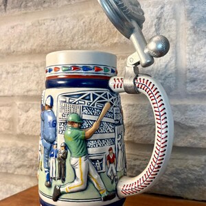 May include: A ceramic beer stein featuring a relief of a baseball game with players in green and blue uniforms. The handle is shaped like a baseball with red stitching, and the metal lid is topped with a baseball glove. The base displays the year 1980.