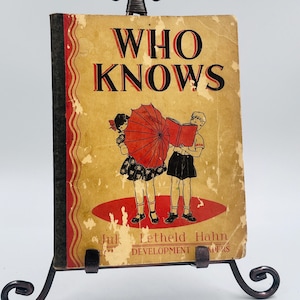 Puede incluir: Libro antiguo titulado "WHO KNOWS" con una cubierta desgastada de color beige. La portada presenta un título rojo y negro, y una ilustración de dos niños, uno sosteniendo un paraguas rojo y el otro leyendo un libro. El libro se exhibe en un soporte de metal decorativo.
