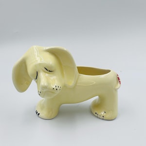 May include: A ceramic dog planter in a light yellow color. The dog has closed eyes, black spots, and a red heart detail on its rear. The planter has an open top for plants or storage.