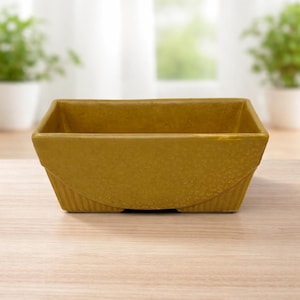 Puede incluir: Una jardinera cuadrada de color mostaza con una superficie texturizada. La jardinera tiene un diseño ligeramente curvado en los lados y es adecuada para uso en interiores o exteriores. La jardinera está hecha de cerámica o un material similar.