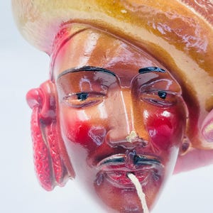 May include: A ceramic figurine of a man's face, featuring a sombrero. The face is painted with reddish-brown and red hues, with black details for the eyes and mustache. A white string extends from the mouth. The hat is tan and yellow.