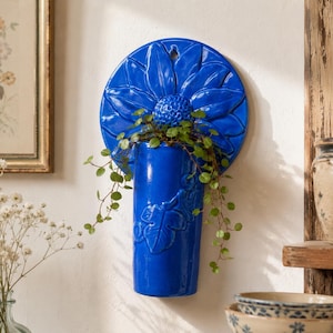 Puede incluir: Una maceta de pared de cerámica en color azul cobalto vibrante con un diseño de girasol en relieve en la placa trasera circular y una base cilíndrica con motivos de hojas. Una pequeña planta verde cuelga de la abertura de la maceta.